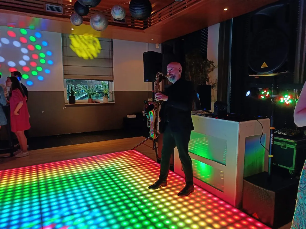 Saxofonist Franklin Schieman speelt live naast de DJ-booth, op een verlichte dansvloer in regenboogkleuren